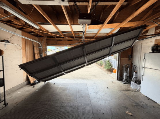 Broken overhead garage door collapsed and dangling diagonally in an open garage position, exposing wooden rafters and garage contents in the background.